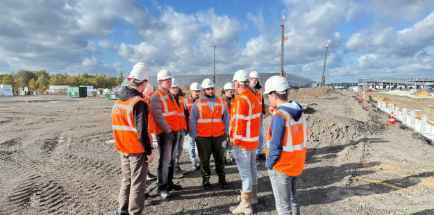Groep studenten op de bouwplaats van Westfields III in Oirschot, waar zij uitleg krijgen over de voortgang van het project en de toegepaste bouwtechnieken.